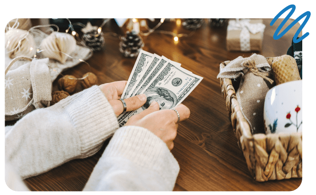 Woman sitting at table surrounded by gift baskets and festive lights, holding $100 bills in hands.