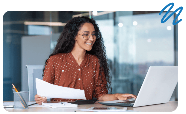 Happy business woman sitting at desk holding documents in one hand and navigating laptop with the other.