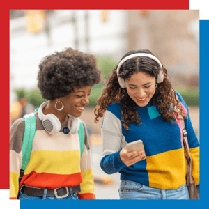 Two women is colorful sweaters walking next to one another with headphones looking at phone screen.