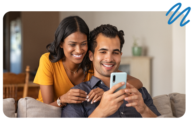 Woman with arms around man while he sits on couch, both looking at cell phone in his hands.
