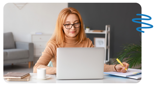 Stylish woman with glasses sitting at desk looking down at laptop screen and holding pen in left hand.