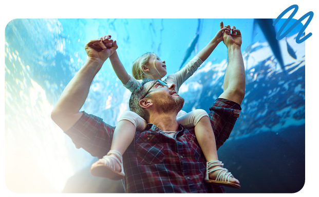 Father holding young daughter and shoulders while both look up at marine life in aquarium tank.