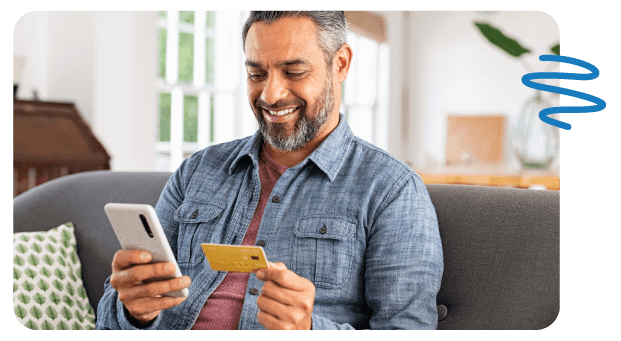 Happy man sitting on couch holding cell phone in one and hand credit card in the other.
