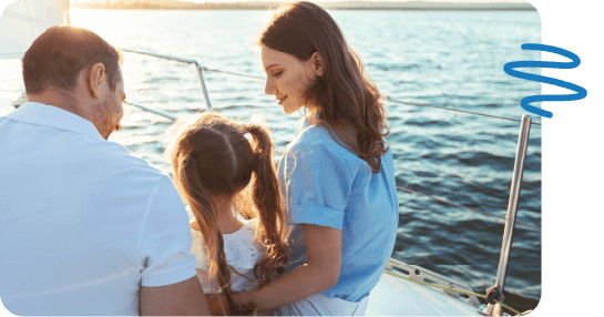 Family of three sitting on deck of boat looking at one another with ocean water beside them.