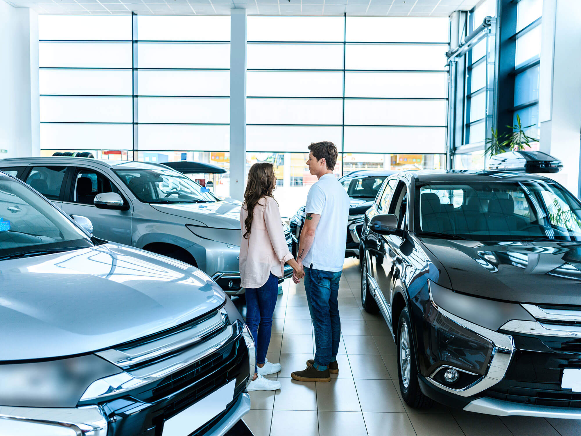 Couple holding hands standing amongst cars in dealership.