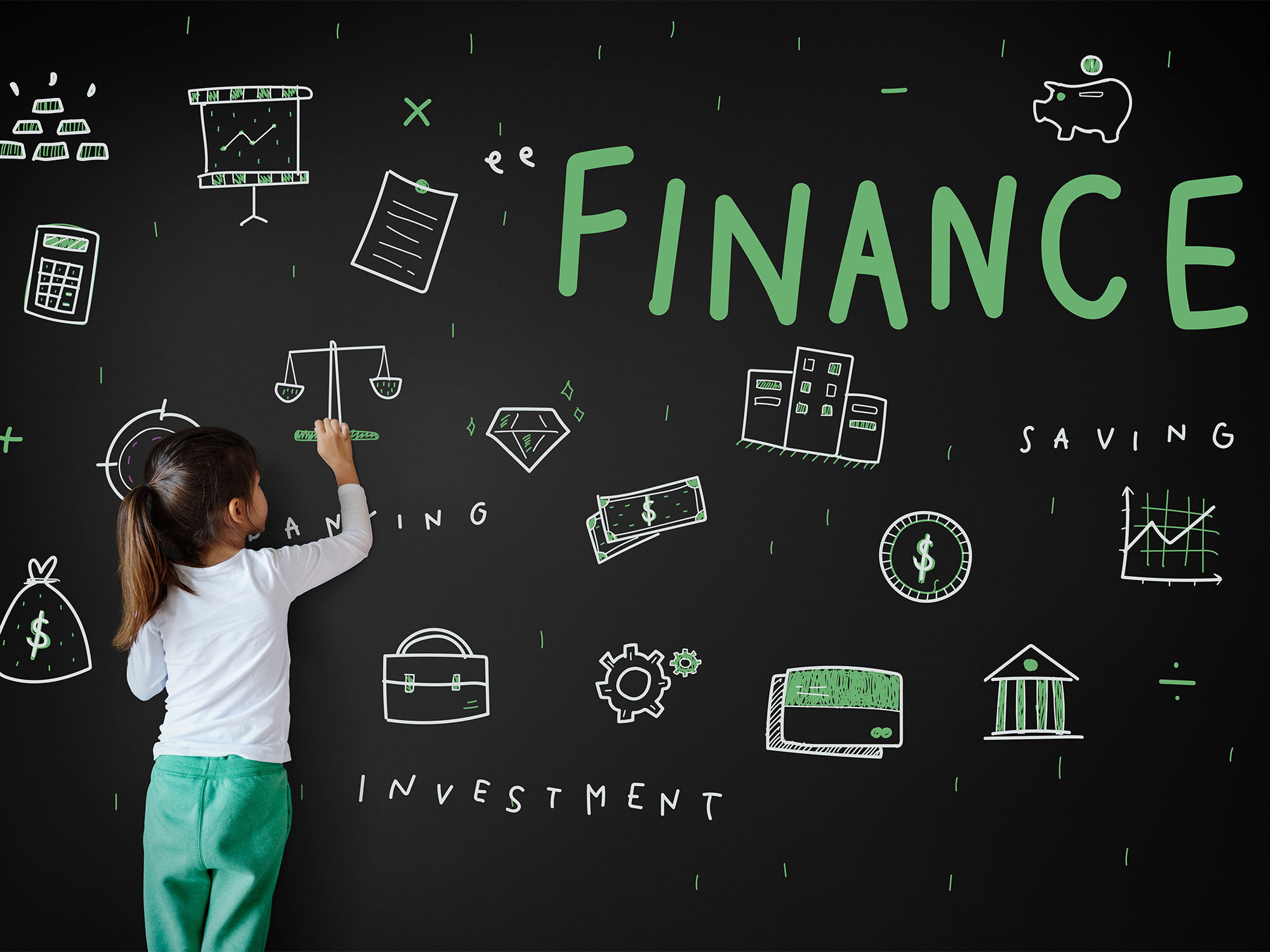 Little girl drawing on chalkboard covered in financial icons and words.
